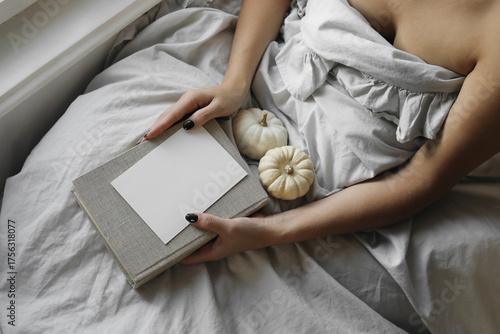 Cozy autumn morning scene. Woman relaxing in bed holding book, blank greeting card invitation mockup. Blanket, small white pumpkins, soft linens. Neutral tones, gentle light. Warm peaceful atmosphere.