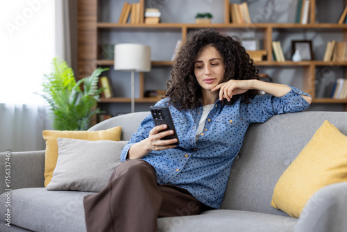 Fototapeta Woman with dark curly hair sitting on a gray sofa in her living room, calmly scr