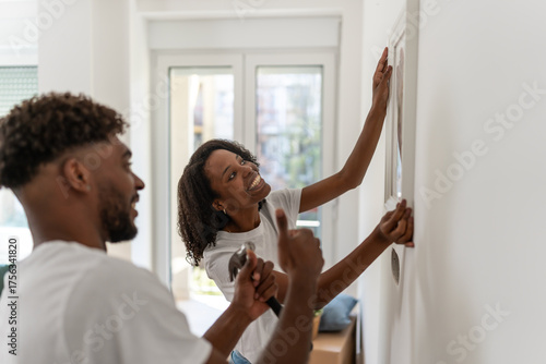 Happy African American couple hanging a picture on the wall while decorating their new home. Smiling and working together, enjoying teamwork, love, and joy in creating a cozy space