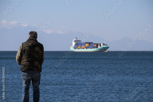 man in camouflage vest gazes at a turquoise container ship on the blue sea, with distant snow-capped mountains under a clear sky, symbolizing travel or commerce