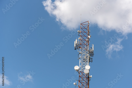 View of a massive telecommunications antenna or transmission tower, supporting various communication antennas for mobile telephony, radio, TV, and internet. The background is a vibrant blue sky.
