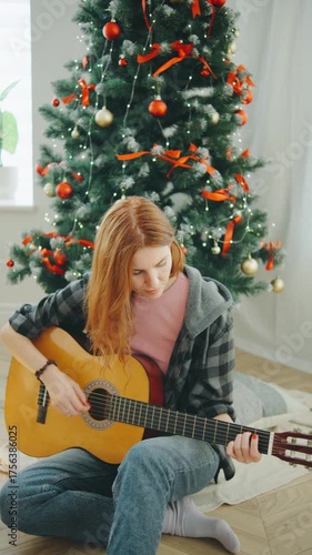Young woman playing guitar in front of Christmas tree, creating a joyful holiday atmosphere at home during winter afternoon