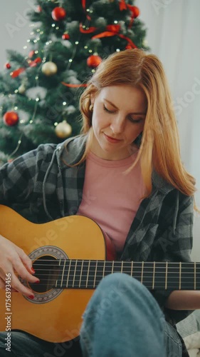 Young woman playing guitar beside a decorated Christmas tree during the holiday season in a cozy indoor setting with festive lights