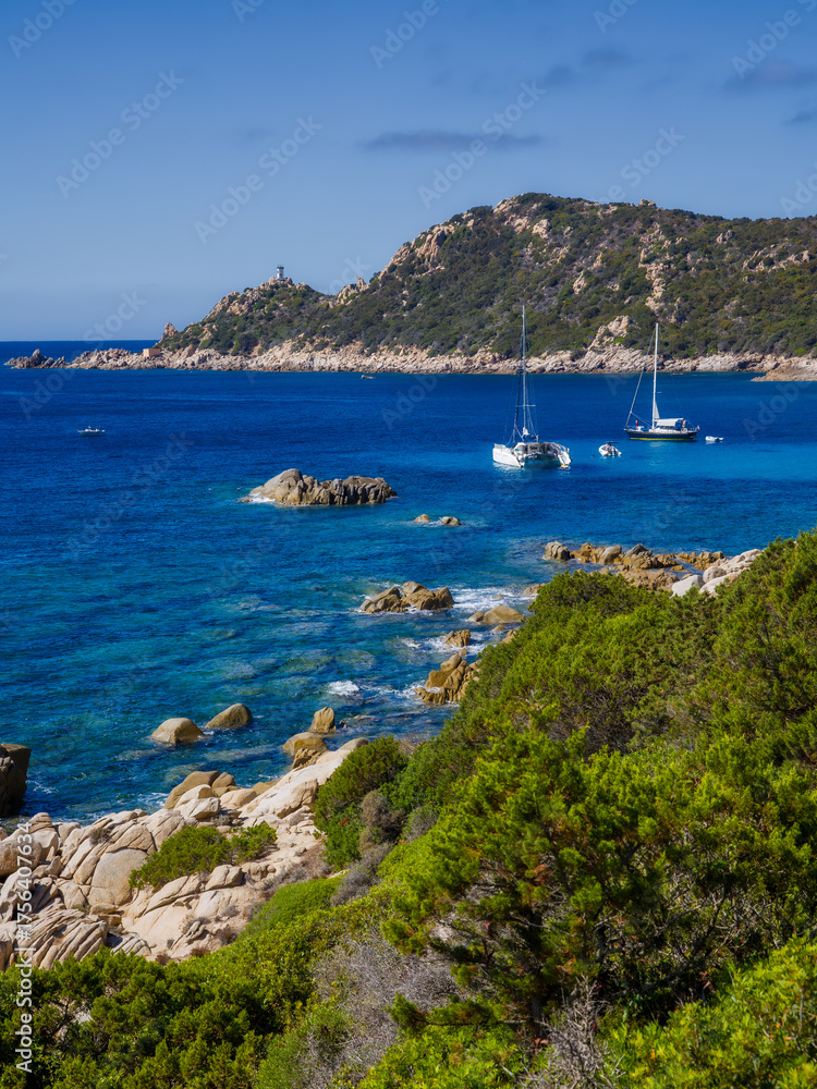 Obraz premium the bay of Cap Muro with its beautiful turquoise water, boats at anchor, and the lighthouse on the hill in sunshine and blue skies