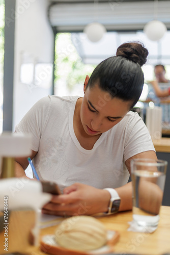 Joven mujer latina concentrada trabajando o estudiando en una cafetería moderna con luz natural, enfocada en sus tareas.