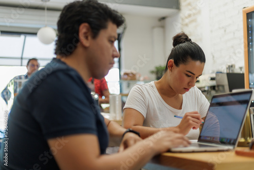 Joven mujer latina concentrada trabajando en su laptop en un café moderno con un hombre en primer plano.