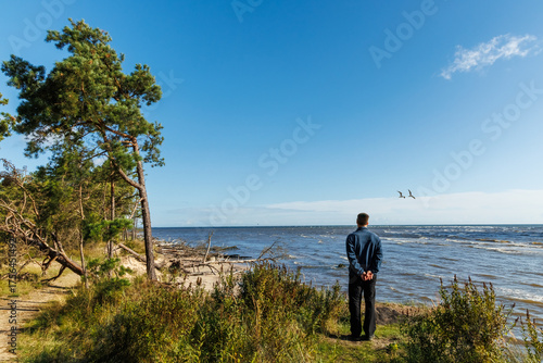 Man standing on a grassy coastal cliff looking at the sea under a clear blue sky, peaceful seaside moment with nature and freedom atmosphere