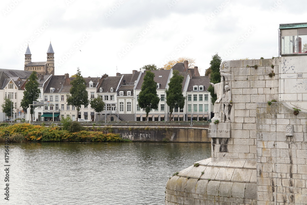 Fototapeta premium Autumn Scene Along Maastricht Riverfront With Historic Buildings And Sculpted Stone Bridge By Calm Waterfront Quays