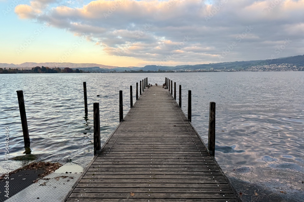 Fototapeta premium Hünenberg am See: A wooden walkway leads out onto Lake Zug, with an autumnal evening atmosphere and clouds in the sky. Symbolic of tranquility, meditation, and peacefulness.