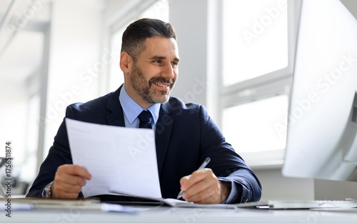 Happy middle aged businessman working with computer and documents in office, entrepreneur in suit sitting at desk, checking company financial reports