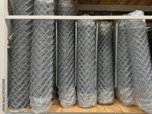 Stacked rolls of galvanized chain link wire mesh, wrapped in plastic, displayed on a hardware store shelf for fencing, security barriers, garden enclosures, and construction projects.