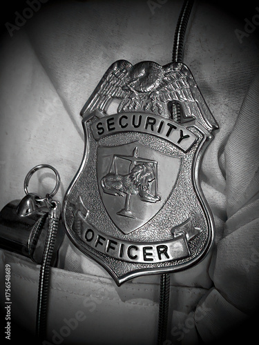 Close-up of a private security officer badge clipped to a white uniform. The badge features an emblem with scales of justice and a lion in the center. Next to the badge is a metallic whistle. 