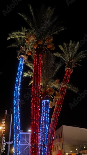 Vertical video: three palm trees wrapped in red and blue holiday lights against dark sky at night in Fethiye, Turkey. Festive New Year and Christmas concept