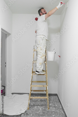 Painter on ladder rolling white paint on wall