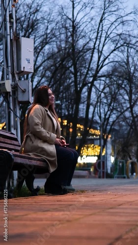 brunette woman wearing coat sitting on the bench in the street at night using smartphone