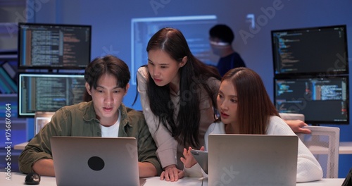 Focused group of young Asian developers working together on a coding project at night. Collaboration around laptops in a modern tech office with code screens and software development teamwork.