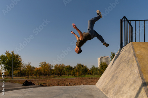 Male freerunner performs a backflip off a sloped concrete wall in an outdoor park, captured midair with extended limbs under clear skies, showcasing athletic precision and agility