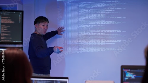 Asian male developer presenting source code to colleagues in a dark tech office. Team listening to coding explanation during software training, AI session, or cybersecurity presentation.