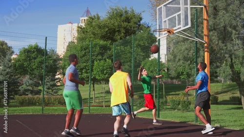 Active sporty fit basketball player making successful assist to African American male teammate, helping to score points with layup shot while multi ethnic sports teams playing game on outdoor court