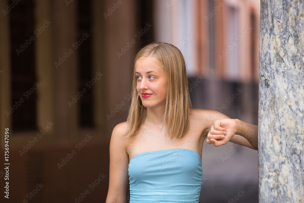 Naklejka premium Young woman, blonde, blue eyes, slim, wearing a light blue dress, innocent and smiling, looking distracted, leaning against a marble column. Concept: femininity, innocence, youth.