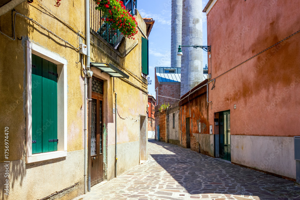 Fototapeta premium Colorful narrow street with traditional buildings, green shutters, and glass factory chimneys on Murano island, Venice, Italy. 