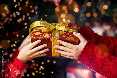 Artistic composition showing the magic of giving at Christmas time. Two pairs of hands gently holding a red gift box with a golden bow against a glowing background with soft bokeh lights. 
