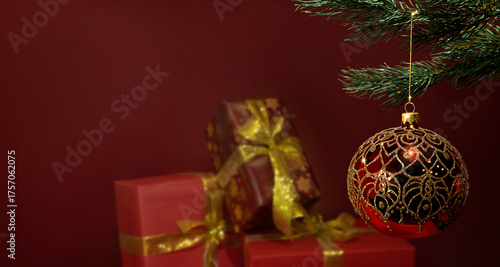 In the foreground is a red Christmas ball with an elaborate gold pattern hanging from a green tree branch. 