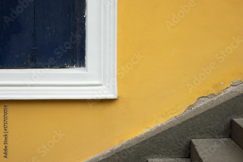 A closed old wooden shutter in an intense cobalt color, the yellow facade of the building, and a fragment of the stairs. Architecture, fragment, color, contrast.