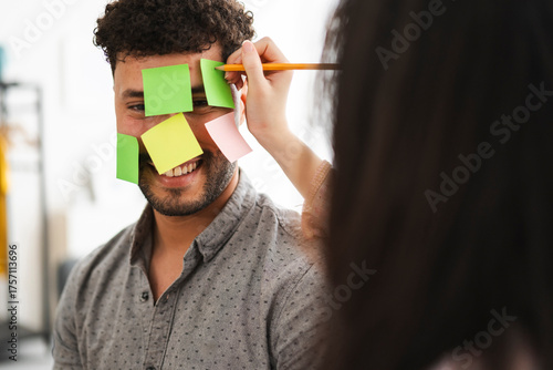 A person writes on sticky notes attached to another person's face, symbolizing brainstorming or a creative session.