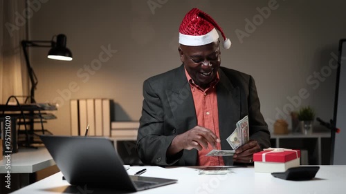 Happy senior African American businessman wearing a Santa hat and counting his Christmas bonus money in the office at night, smiling and planning his holiday expenses and gifts
