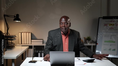 Senior African American businessman sitting at his desk at night, actively participating in a video conference on his laptop, gesturing and speaking passionately while working late in the office
