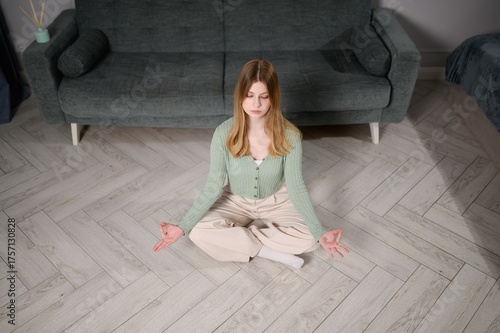 Young woman practices yoga, meditation at home, sitting on living room floor in lotus position, relaxing with eyes closed. Relaxed young woman practices Padmasana yoga pose at home. Relaxation, yoga