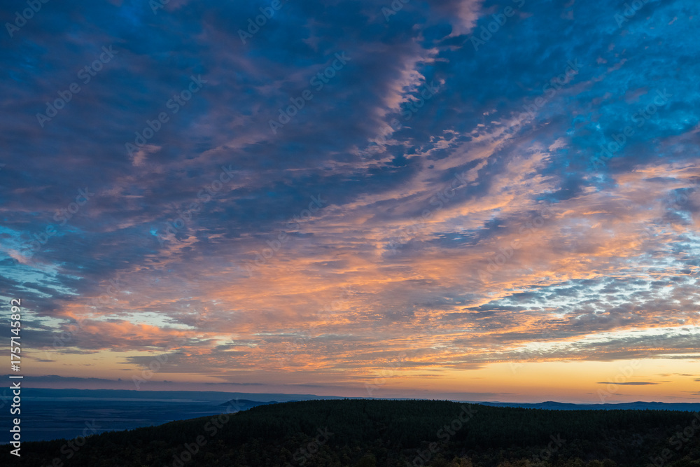Obraz premium A vibrant sky of blue and orange glows above the dark silhouette of forested hills during a tranquil sunrise or sunset.