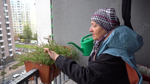 An elderly woman waters flowers on the balcony from a green watering can. A grandmother takes care of a flower in a pot. Gardening at home