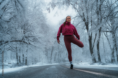 Woman standing on a snowy road, stretching her quadriceps against a backdrop of frost covered trees, actively engaging in fitness and exercise during cold weather conditions