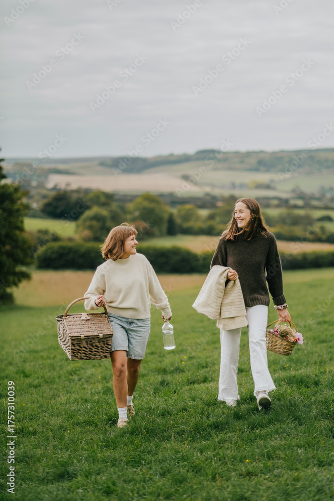 Fototapeta premium Two young women are walking in a field, one of them carrying a basket. The other woman is carrying a bottle of water. The scene is peaceful and serene, with the women enjoying the outdoors