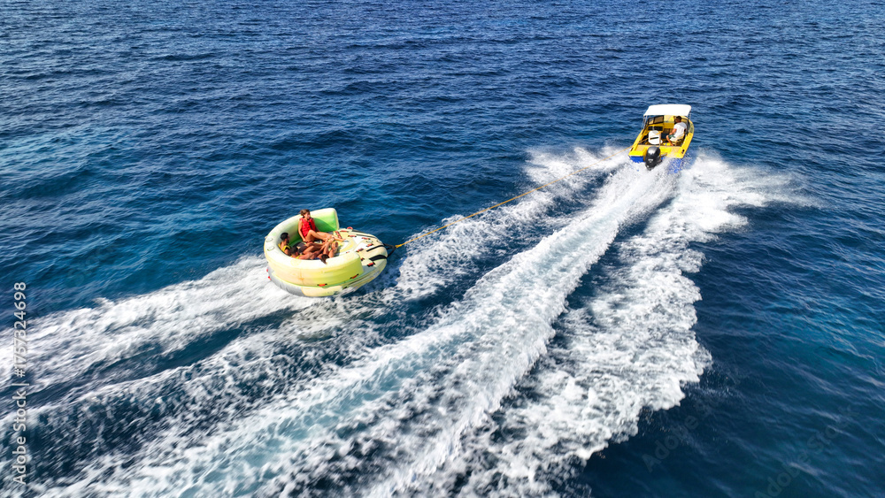 Fototapeta premium Aerial drone tracking photo of powerboat towing inflatable tubes used for water sports activities in deep blue open ocean island sea