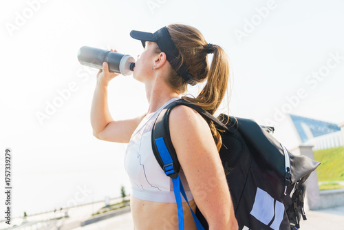 Woman takes a break to hydrate while exercising outdoors near a body of water, showcasing an active lifestyle during a warm day
