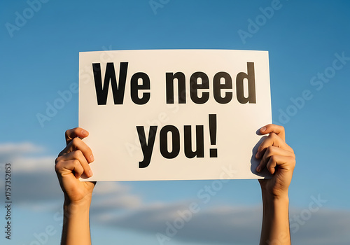 Hands Holding 'We Need You!' Sign Against Blue Sky