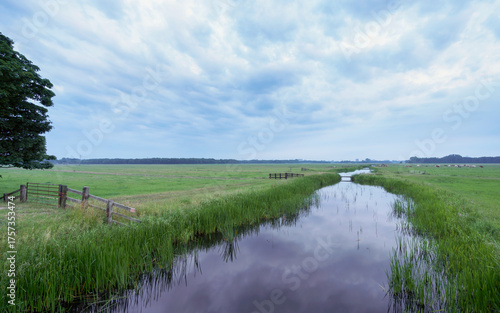 Canvastavla Calm water reflects the cloudy sky over fields
