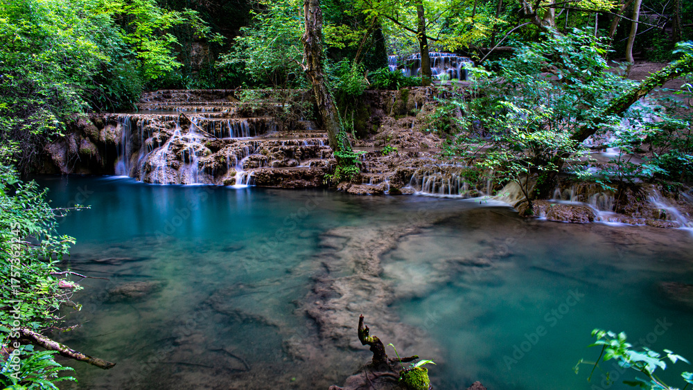 Naklejka premium Long exposure of Krushunski Waterfalls cascading into a turquoise pool surrounded by dense green forest. A serene summer landscape with smooth flowing water and lush vegetation.