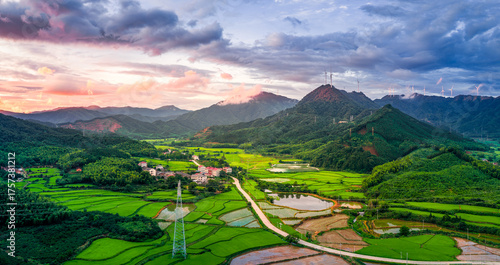 Canvas Print Aerial view of a beautiful rural landscape with a small village, green rice fields, and mountains with wind turbines at sunset