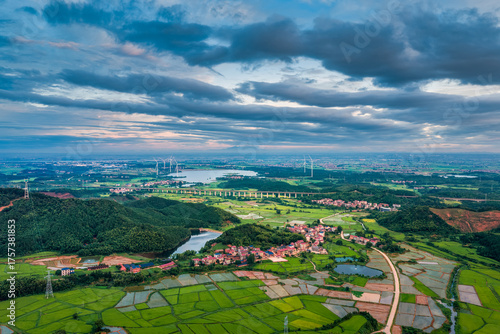 Fototapeta Naklejka Na Ścianę i Meble -  High altitude view of a vast rural landscape with green fields, villages, a lake, and a wind farm under a dramatic sky.