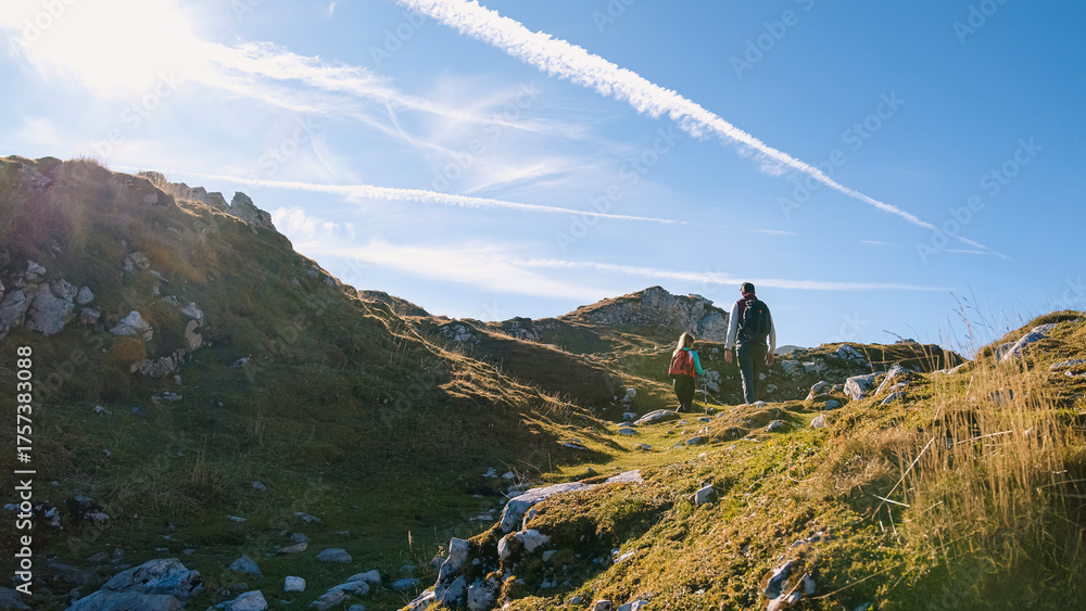 Obraz premium Two hikers traverse a rocky path in the mountains during a sunny day, enjoying the serene landscape and fresh air.