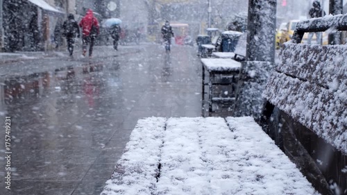 Snowflakes are falling on a snowy bench and blurry people walking at the background in slow motion