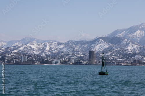Green navigation buoy in sea with snow covered mountains and coastal city view