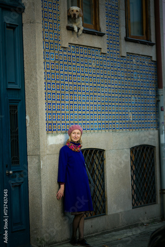 A woman in a lilac coat and beret stands by a wall covered with azulejos in Porto, Portugal, while a dog looks down from the window above. The scene feels warm, poetic, and deeply local.