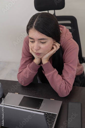 Tableau sur toile A young professional woman stares at her laptop screen with a bored expression, feeling disengaged and tired while completing office tasks in a modern corporate workspace