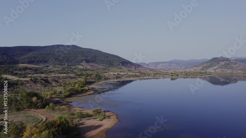 Aerial drone shot rising over the red rocky shoreline of Salagou Lake, revealing blue waters and hilly terrain. Check my portfolio for more Salagou Lake footage.
