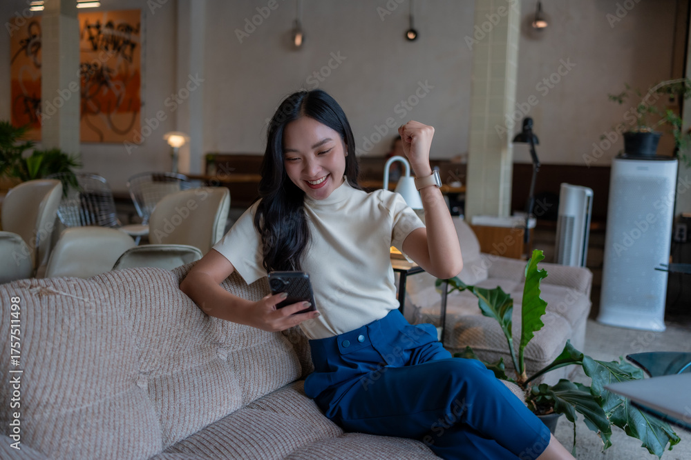 Obraz premium Asian woman celebrating success using smartphone in cafe